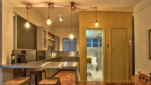A modern kitchen with a stainless steel island, wooden stools, and hanging light bulbs. - Langholm (Cape Town)