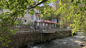 Een stenen brug over een stromende rivier, een houten terras met een pannendak, en een gebouw met meerdere ramen en balkons op de achtergrond.