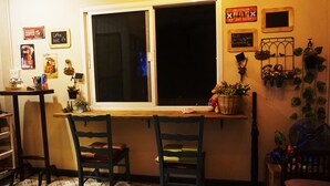 A cozy room with a wooden table and two chairs, a window with a view, and wall decorations including signs and plants. - Staying Hostel (Bang Phli)