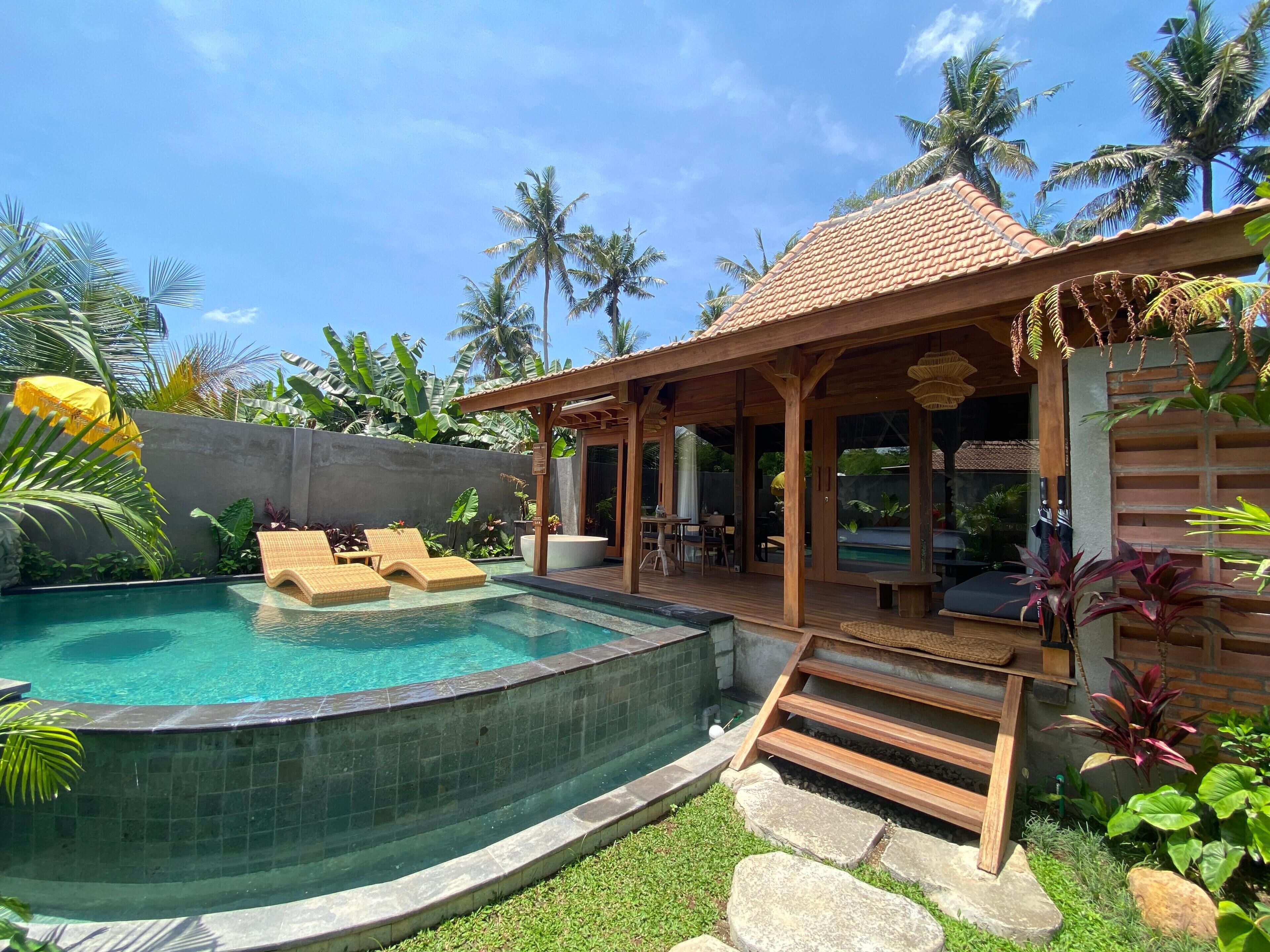 A pool area with lounge chairs, a covered seating area, and tropical plants.
