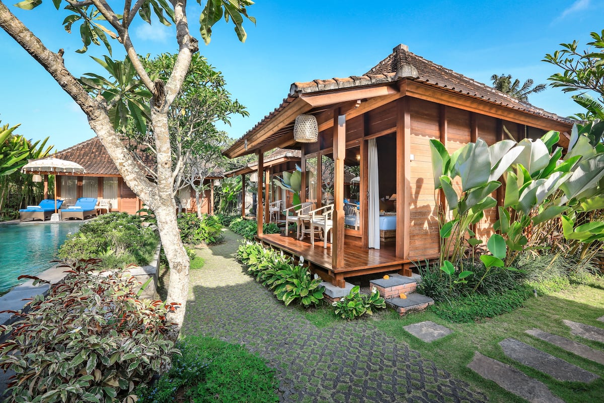 Una casa de madera con balcón, rodeada de abundante vegetación y una piscina.