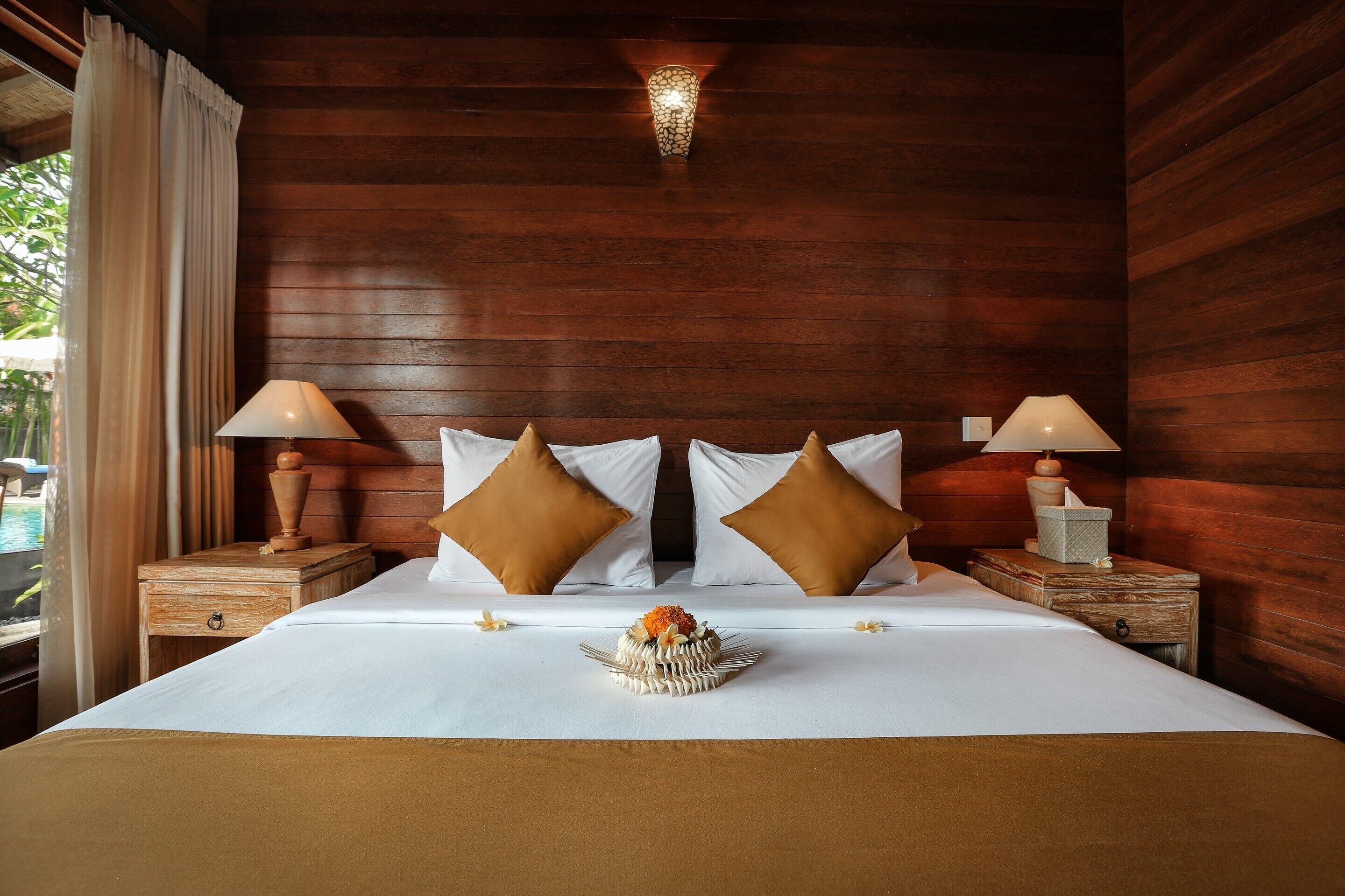 Quarto de hotel com parede de madeira, duas mesas de cabeceira com luminárias, uma cama com roupa de cama branca e duas almofadas castanhas, e uma peça decorativa sobre a cama.