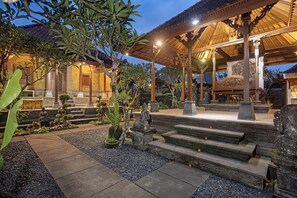 Un pavillon traditionnel en bois, accessible par des marches de pierre, est entouré d’un jardin où poussent différentes espèces de plantes et où se trouve une statue de Bouddha.