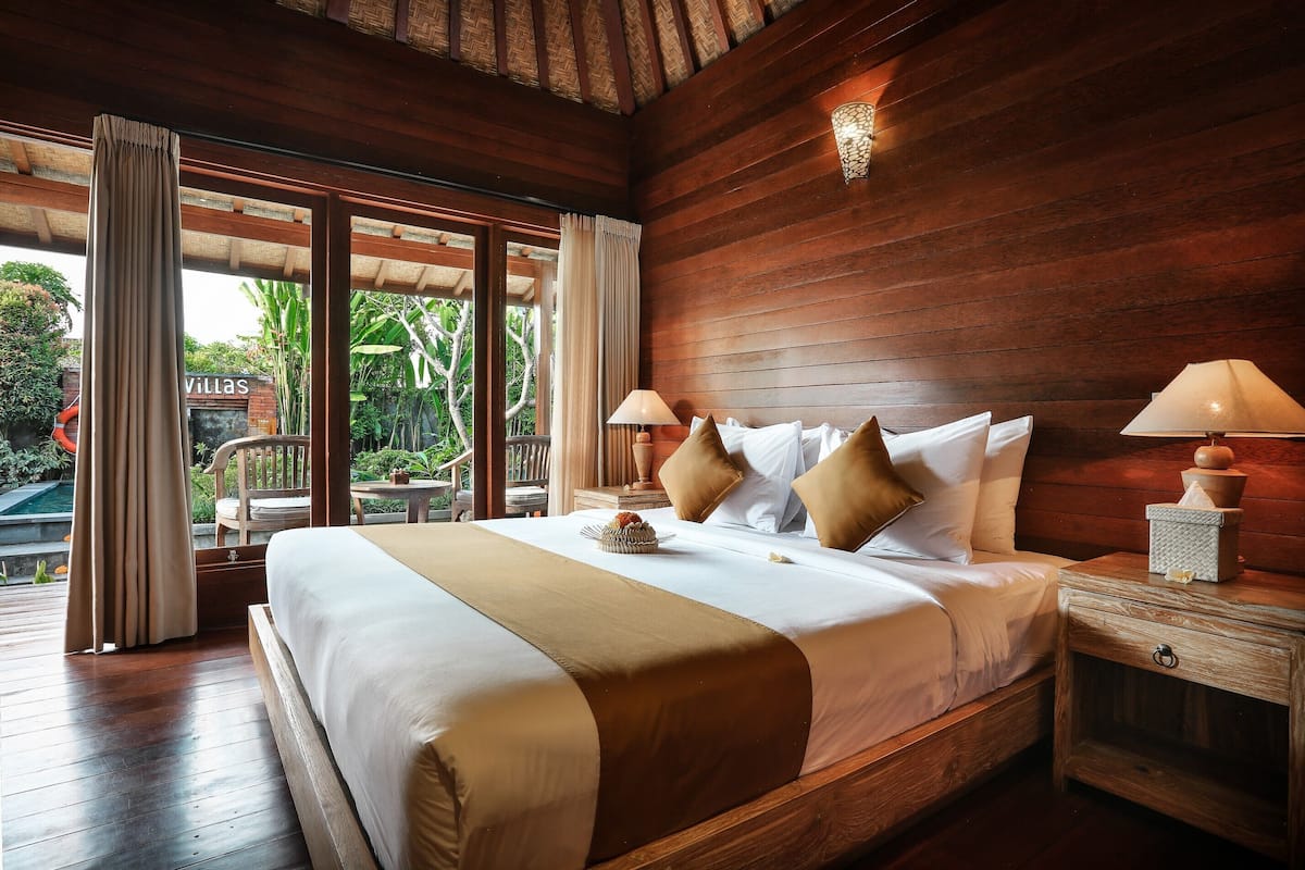 Habitación con paneles de madera, una cama grande, dos lámparas de noche y vistas a una piscina y follaje exterior.