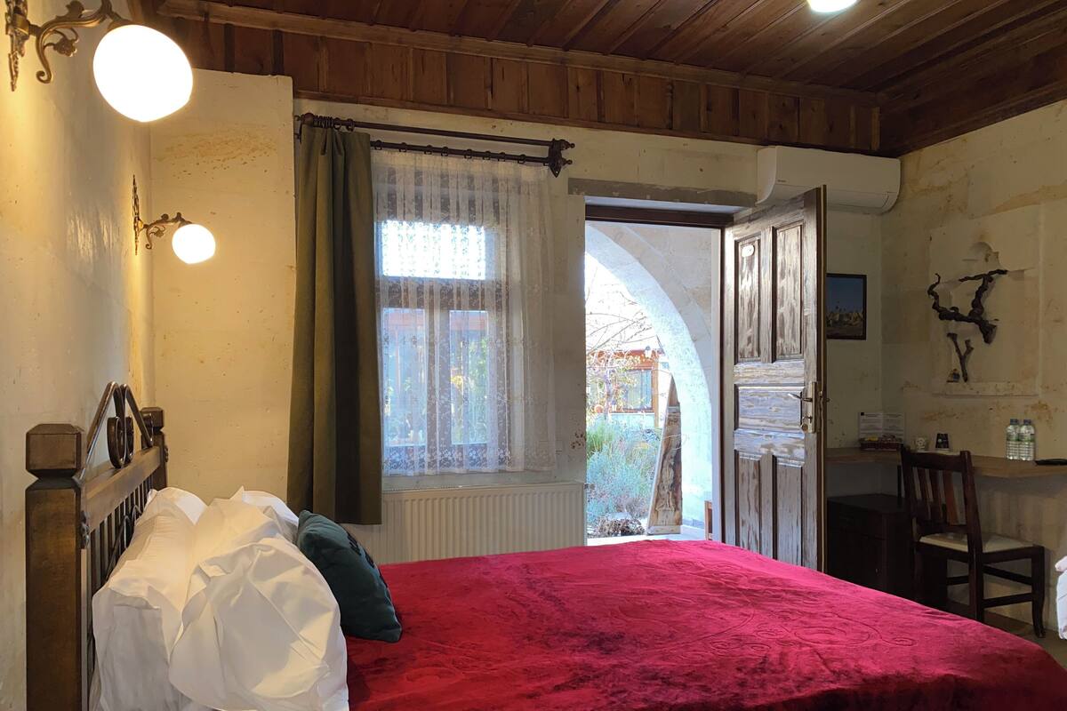 A bedroom with a red bedspread, a wooden headboard, and a window with sheer curtains.