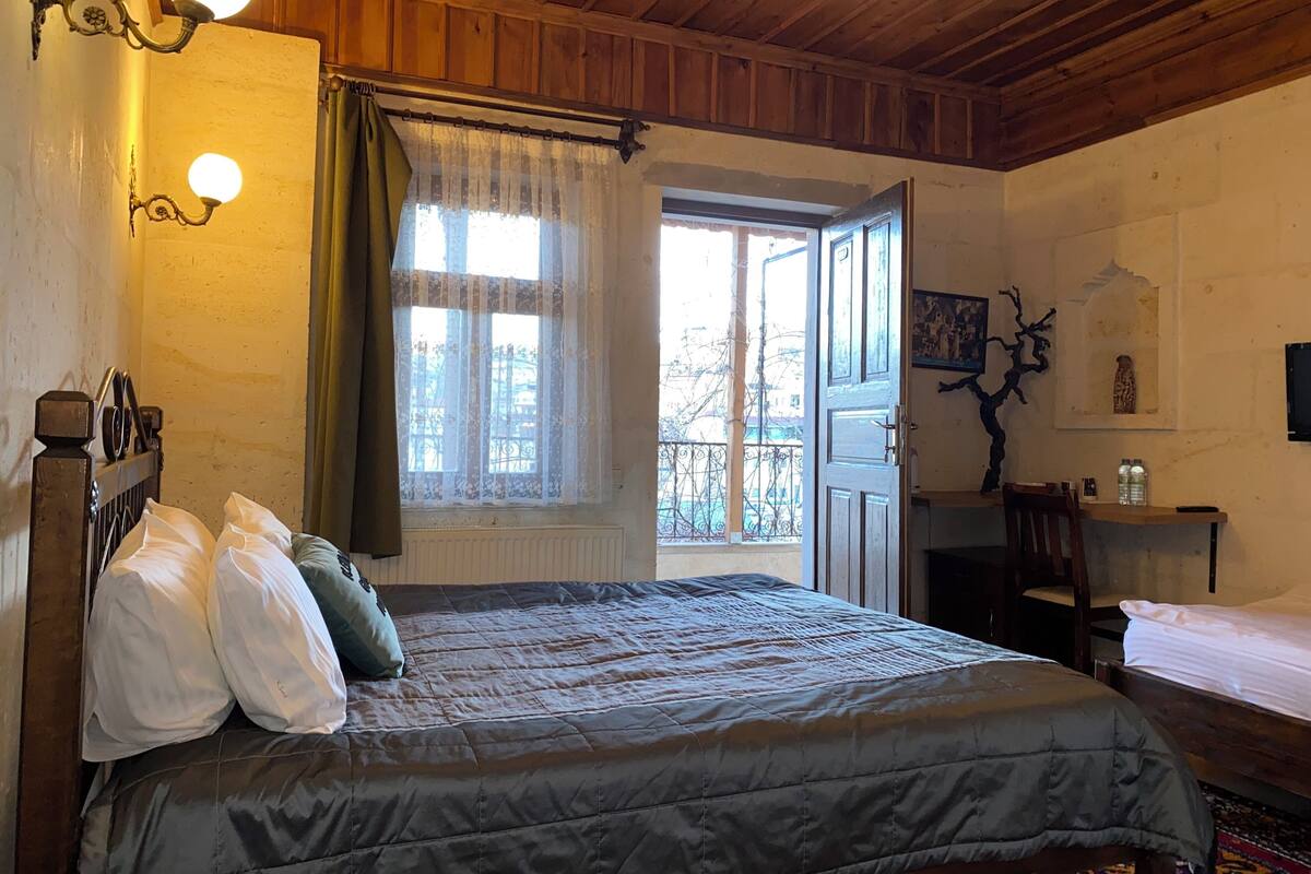 A bedroom with a large bed, a window with curtains, a wooden ceiling, and a small table with a chair.