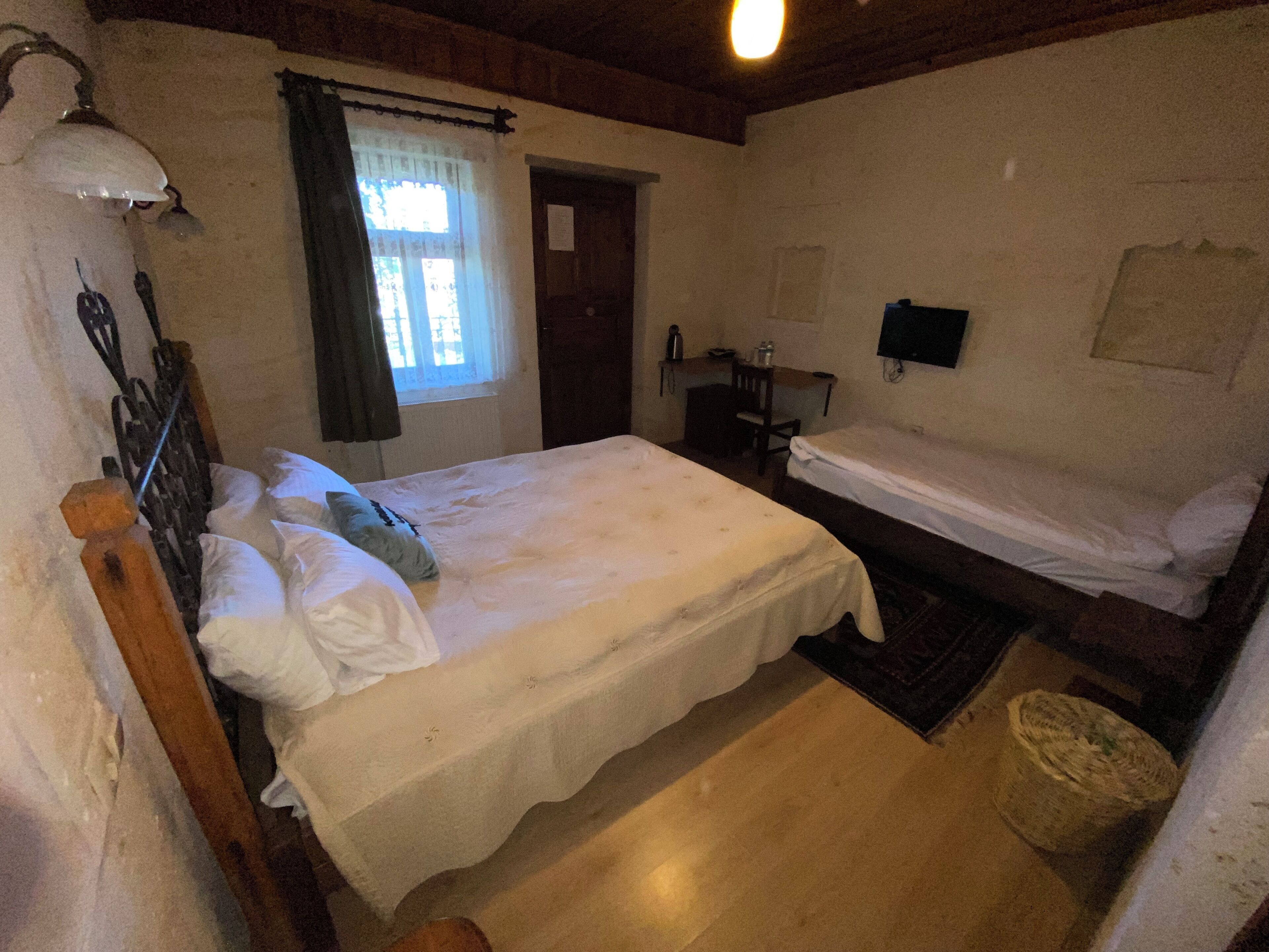 Habitación con dos camas, cabecero de madera, una mesita con una silla, televisor de pared y ventana con cortinas.