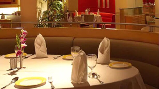 A dining area with a round table set for two, a vase with flowers, and a red curtain in the background. - The Royal Orchid (Mumbai)