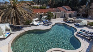 A swimming pool with clear water, surrounded by lounge chairs and umbrellas, set against a backdrop of stone buildings and palm trees. - Villa Diamond (Zupa dubrovacka)