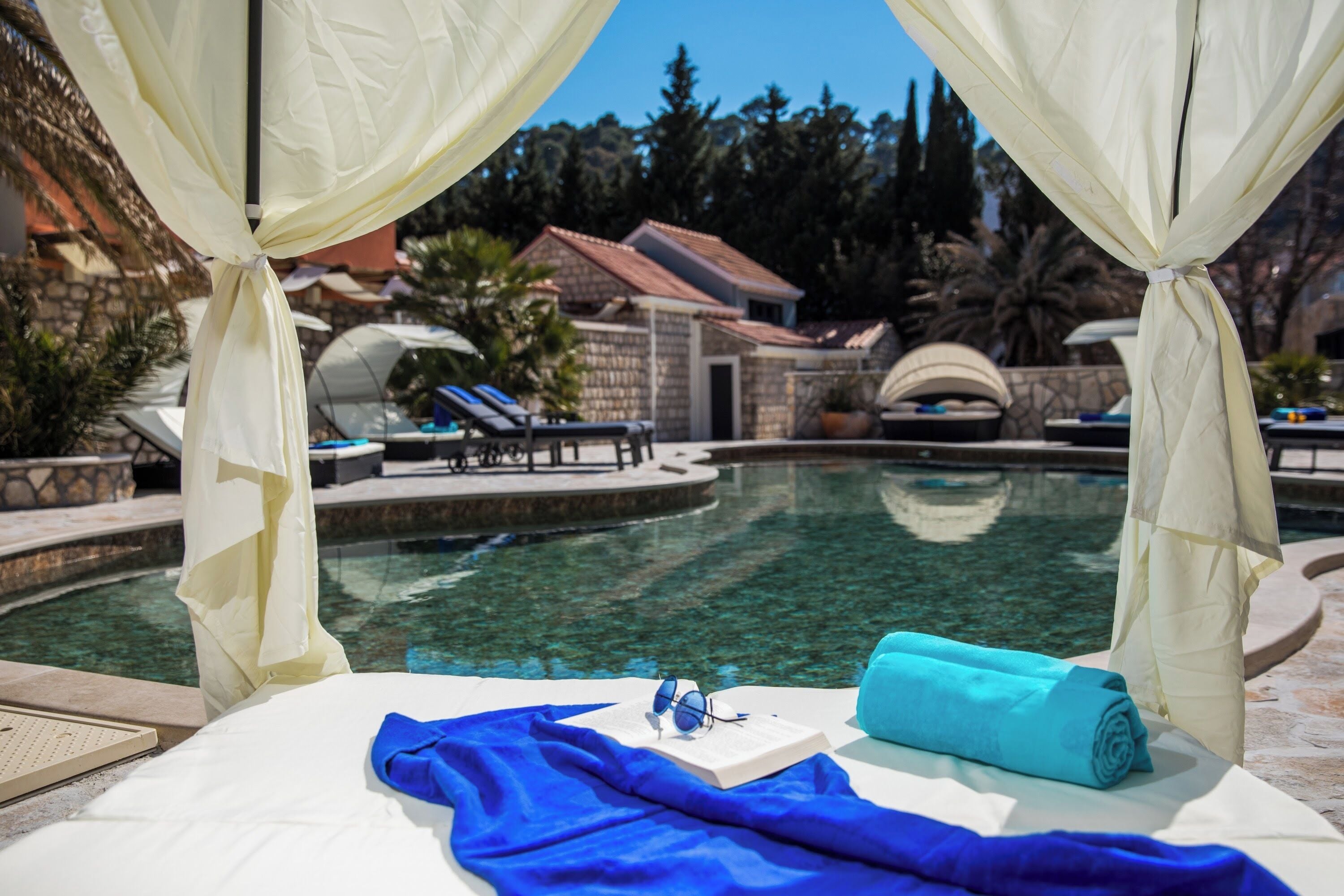 A poolside bed with a book, sunglasses, and a towel, surrounded by a clear pool and a view of a resort area.
