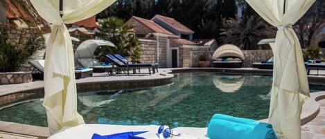 A poolside bed with a book, sunglasses, and a towel, surrounded by a clear pool and a view of a resort area.