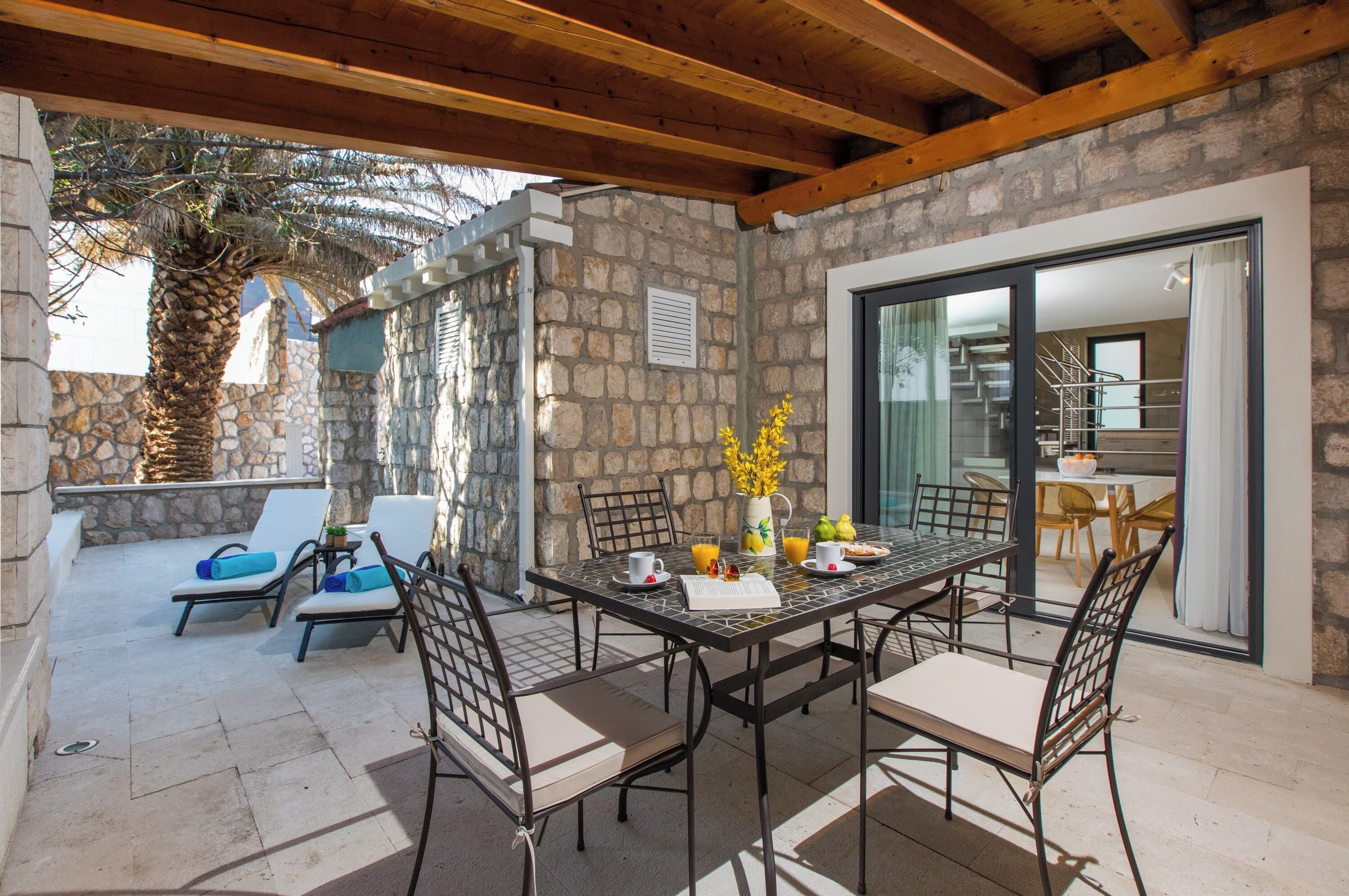 A covered patio with a stone wall, a dining table with chairs, and a view of the interior through a glass door.