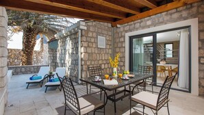 A covered patio with a stone wall, a dining table with chairs, and a view of the interior through a glass door. - Villa Diamond (Zupa dubrovacka)