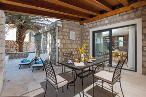 A covered patio with a stone wall, a dining table with chairs, and a view of the interior through a glass door. - Villa Diamond (Zupa dubrovacka)