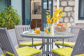 A round table with a glass top, surrounded by four chairs with yellow cushions, a white mug, a glass of orange juice, a vase with yellow flowers, and a basket of fruit. - Villa Diamond (Zupa dubrovacka)