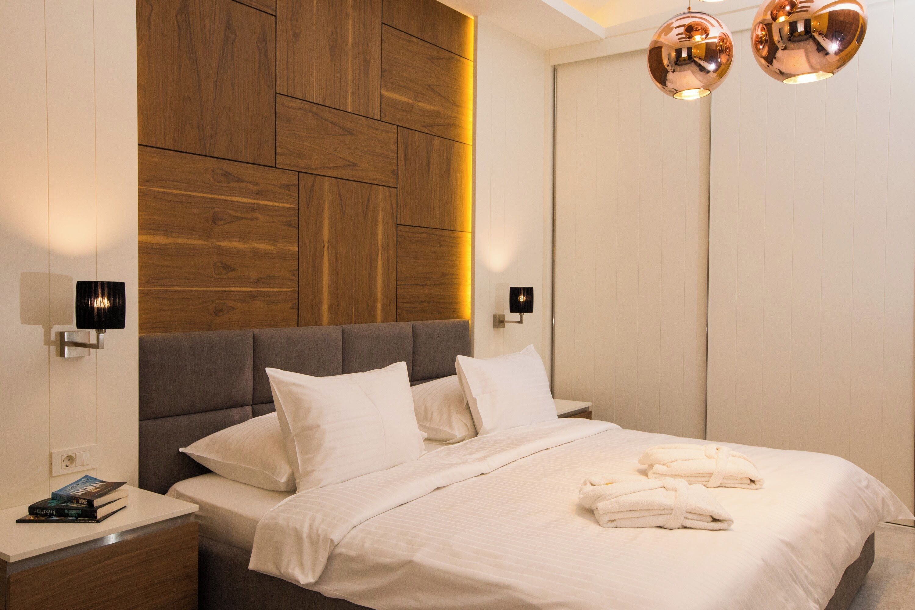 Une chambre moderne avec une tête de lit en bois, un lit recouvert de draps blancs, deux lampes de chevet et une paire de serviettes posées sur le lit.