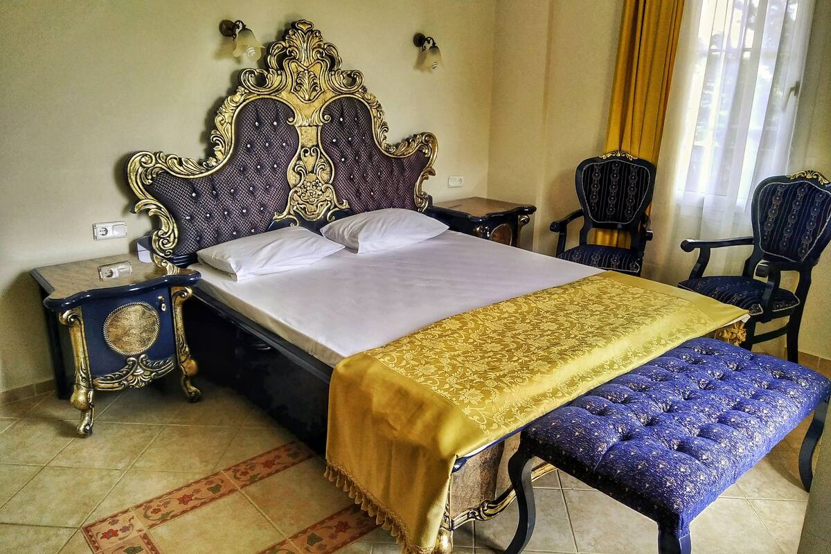 Une chambre à coucher avec un grand lit, une table de chevet, un banc, deux chaises et une fenêtre avec des rideaux.