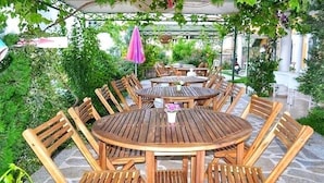 Un área de estar al aire libre con mesas y sillas redondas, rodeada de follaje y una estructura de pérgola.