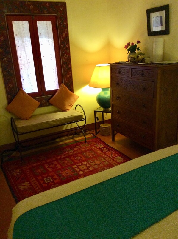 Una habitación con una cama, un banco con cojines, un aparador de madera, una lámpara y una alfombra.