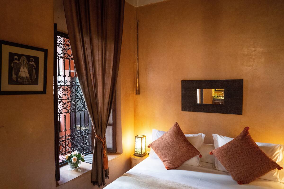 A hotel room with a bed, a nightstand, a wall-mounted mirror, and a window with curtains.