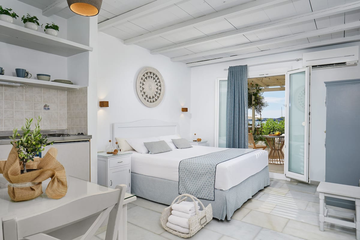 A modern bedroom with a large bed, bedside tables, a small kitchen area, and a balcony with a view.
