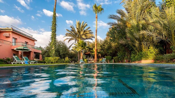 A swimming pool with clear blue water, surrounded by palm trees and a building with a balcony. - Villa Des 3 Golfs (Marrakech)