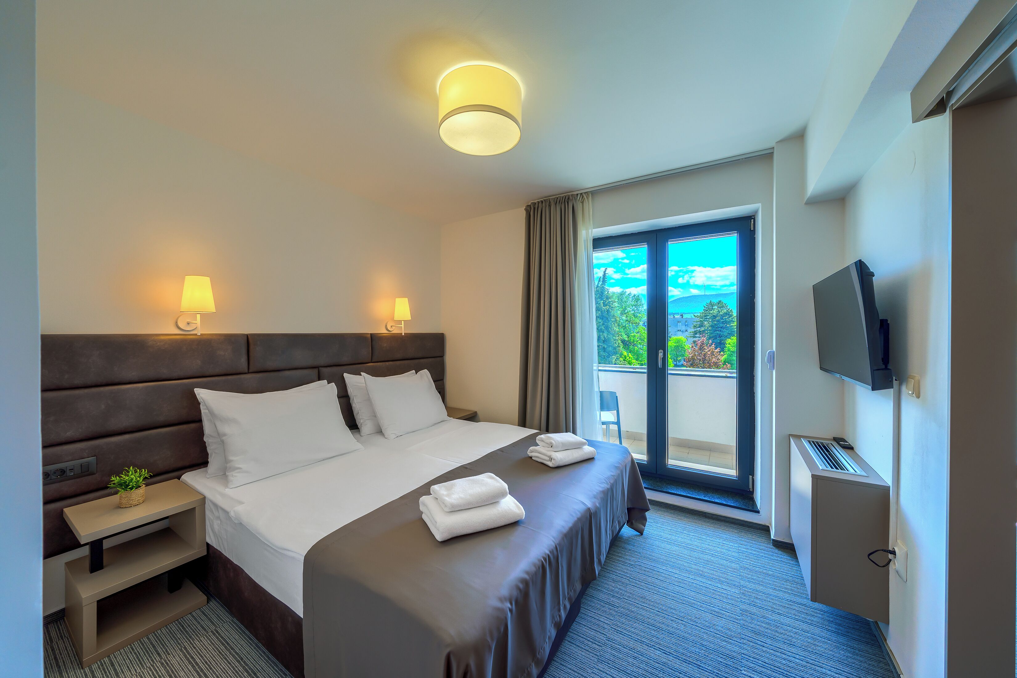 Quarto de hotel com cama, abajures, televisão, uma pequena planta e vista para o exterior.