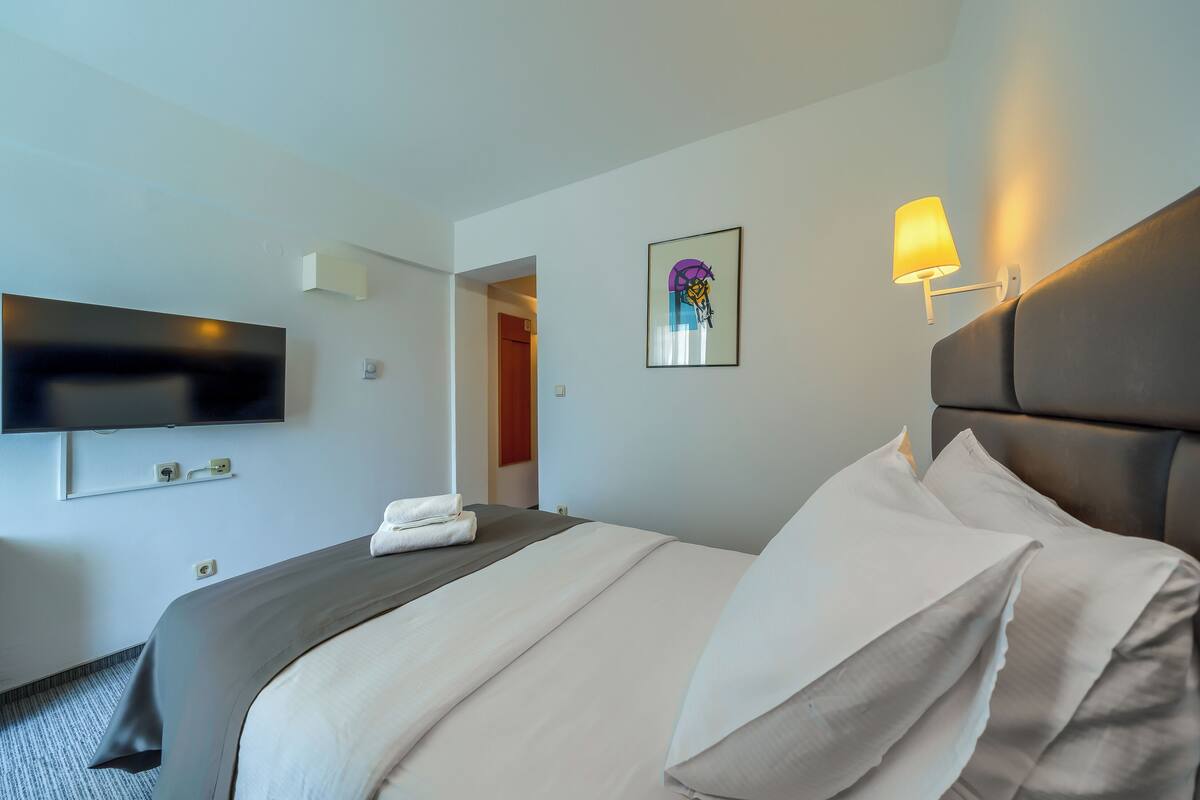 Una habitación de hotel con cama, televisión de pantalla plana, lámpara de pared y una obra de arte enmarco.