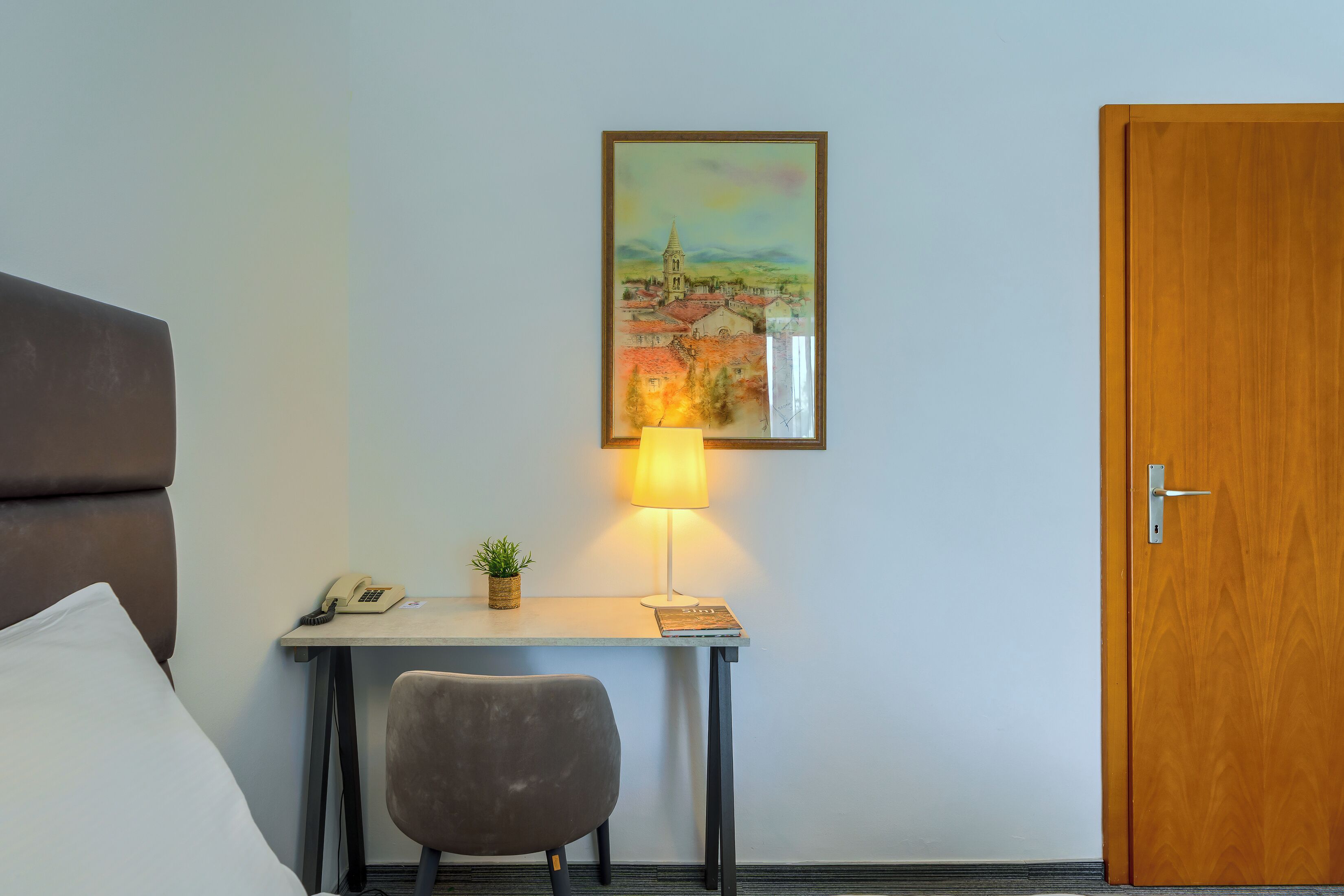 A hotel room with a bed, a desk with a lamp, a chair, a phone, a small plant, and a door.