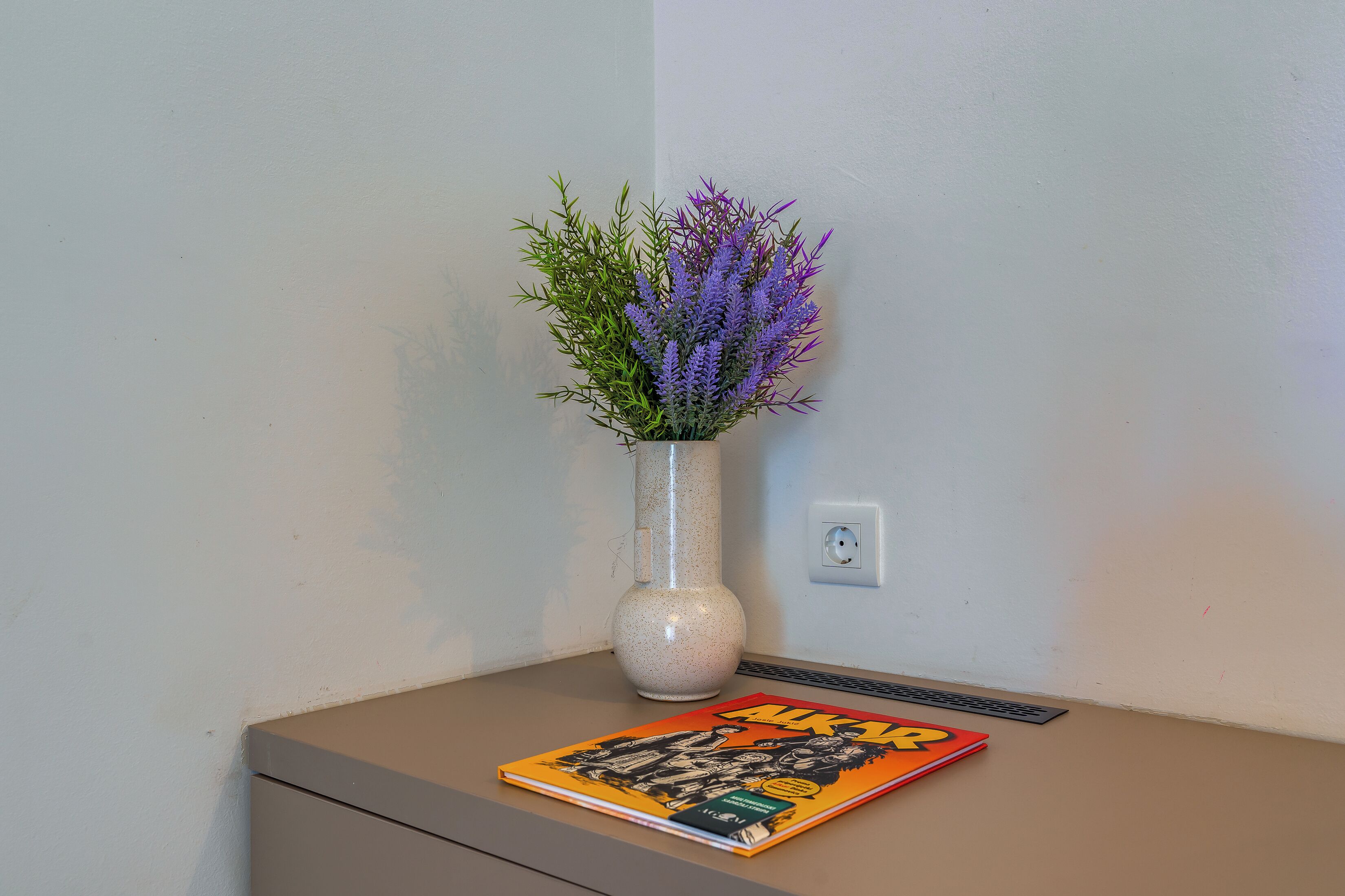 A corner of a room with a vase of lavender flowers on a table, a book, and a power outlet on the wall.