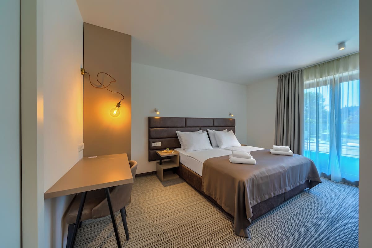 Habitación de hotel con cama, mesita de noche, lámpara y vistas al exterior.