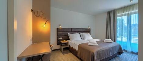 A hotel room with a bed, bedside table, lamp, and a view of the outdoors.
