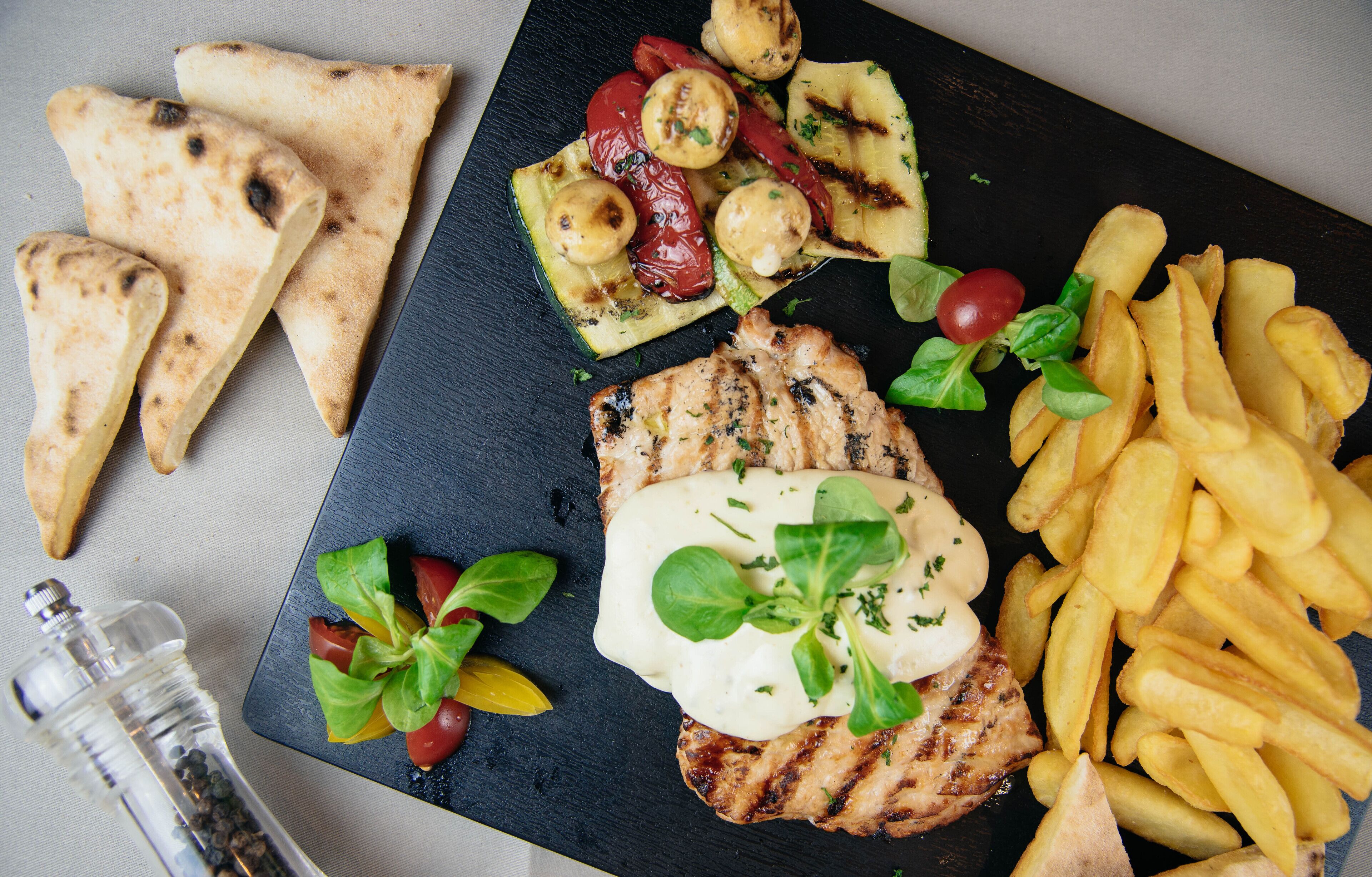 A meal with grilled chicken, French fries, grilled vegetables, and flatbread on a black slate plate.