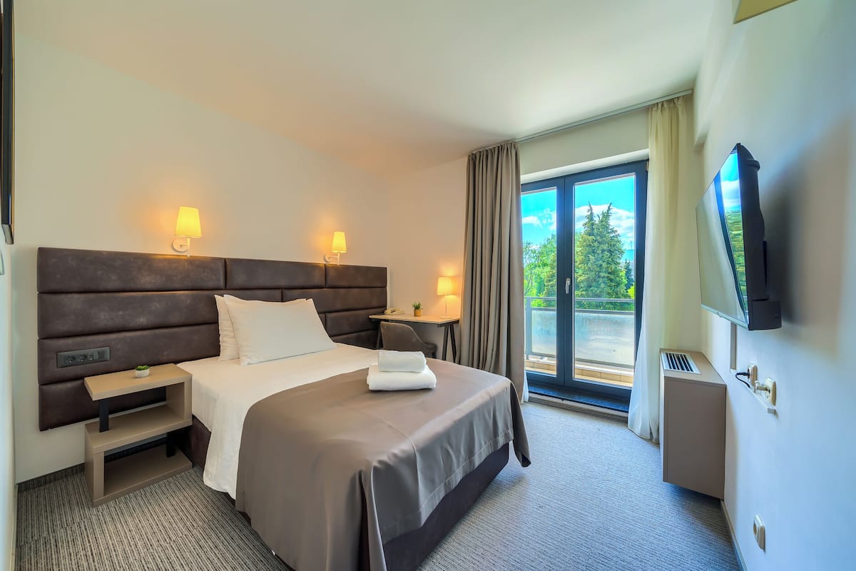 Habitación de hotel con cama, lámparas de noche, televisión, escritorio y vista a zonas verdes por la ventana.