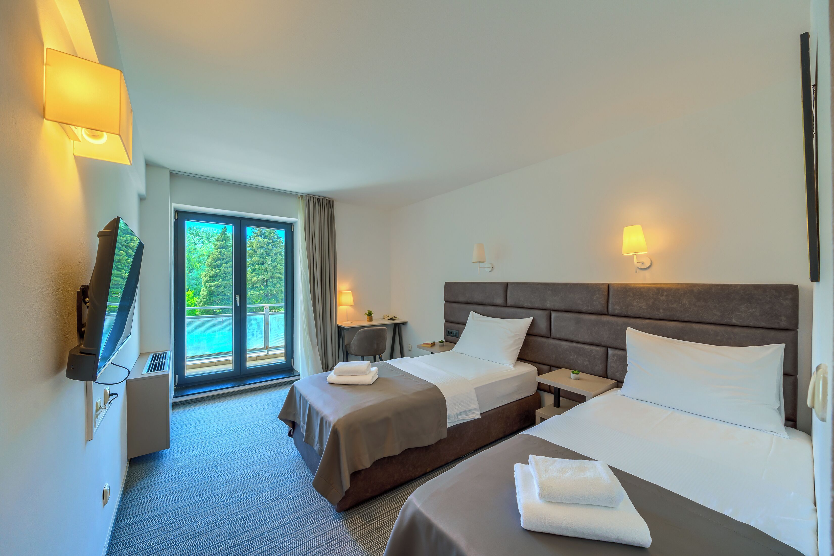A hotel room with two beds, a TV, a desk, and a view of the outdoors.
