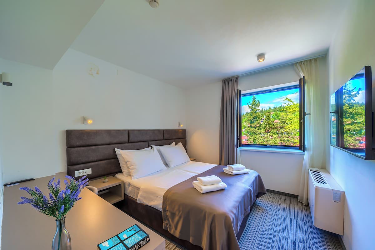 Habitación de hotel con cama, escritorio, florero con flores, televisor de pantalla plana y ventana con vista a vegetación.