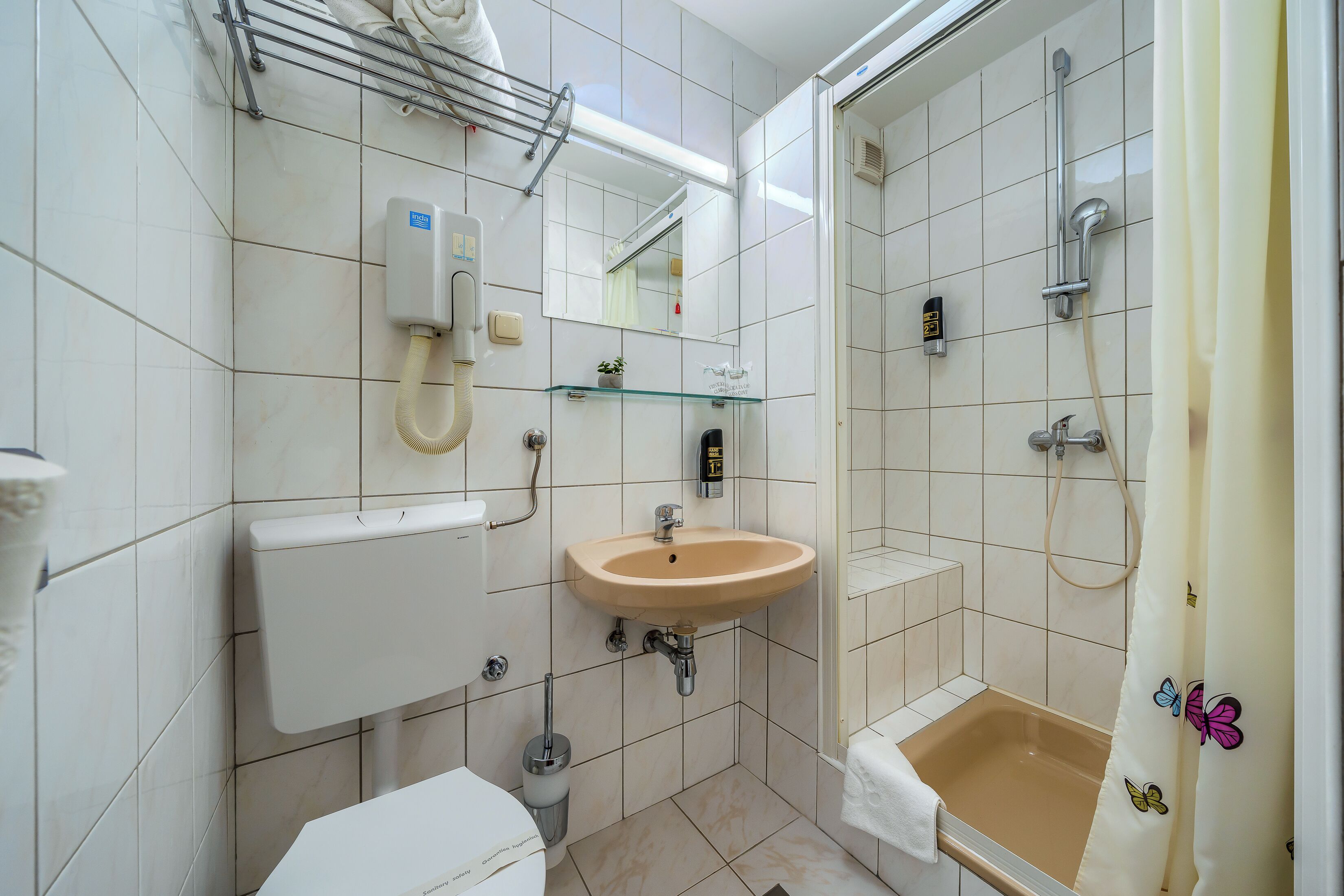A bathroom with a toilet, sink, mirror, shower, and a glass shelf.