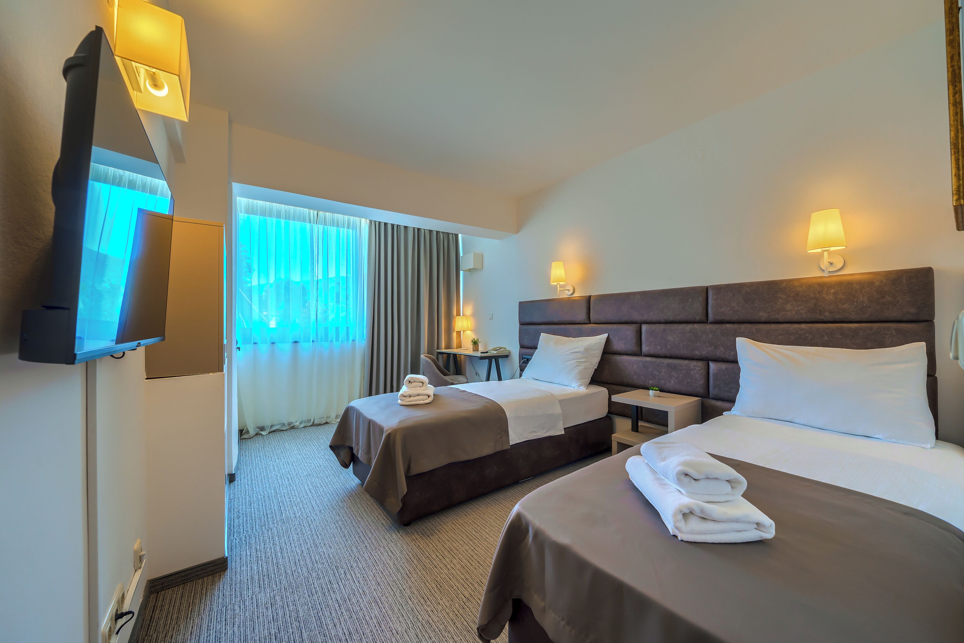 A hotel room with two beds, a TV, bedside lamps, a desk, and a window with curtains.