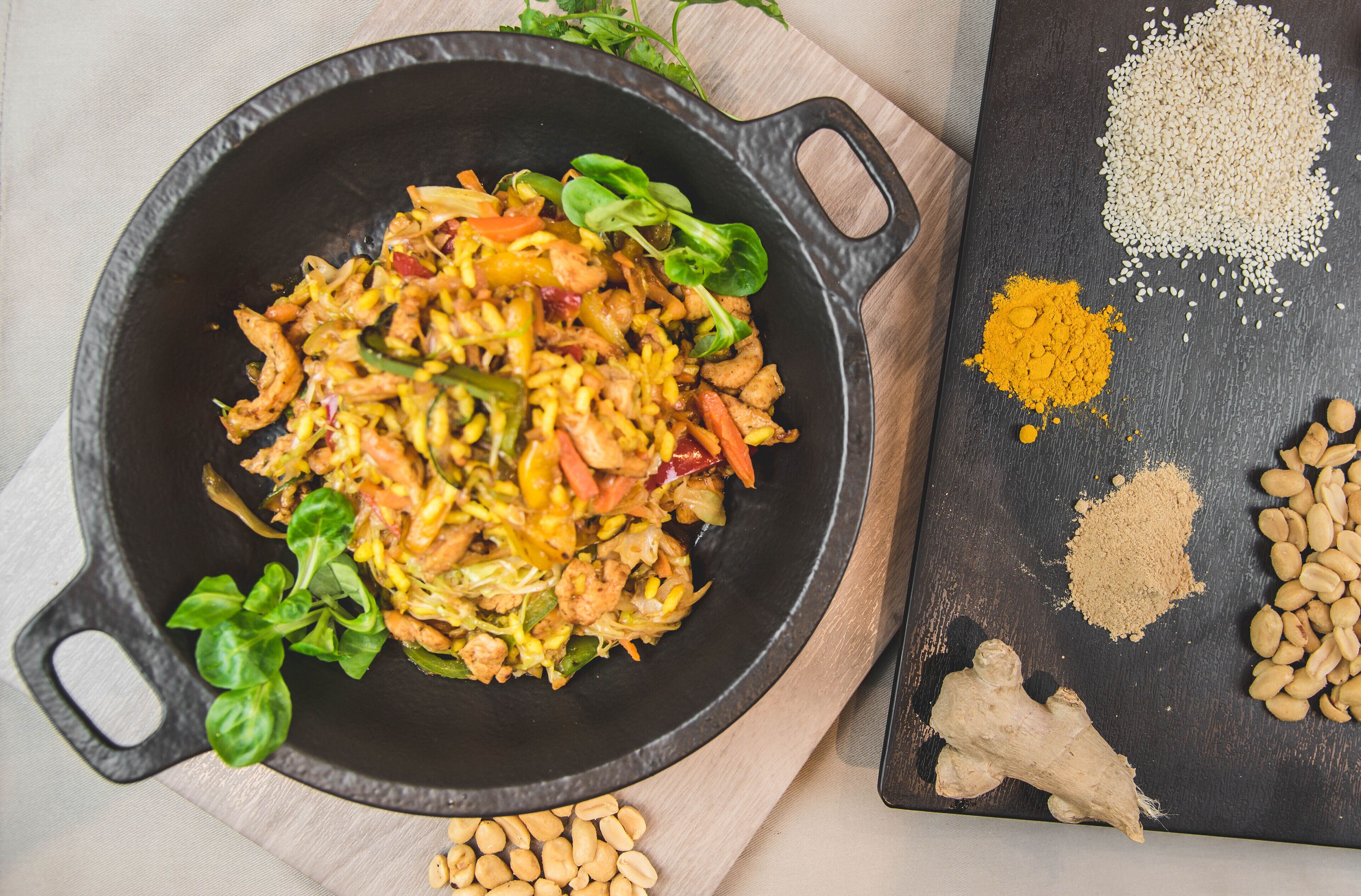 A skillet with a stir-fry dish, surrounded by various spices and ingredients on a wooden board.