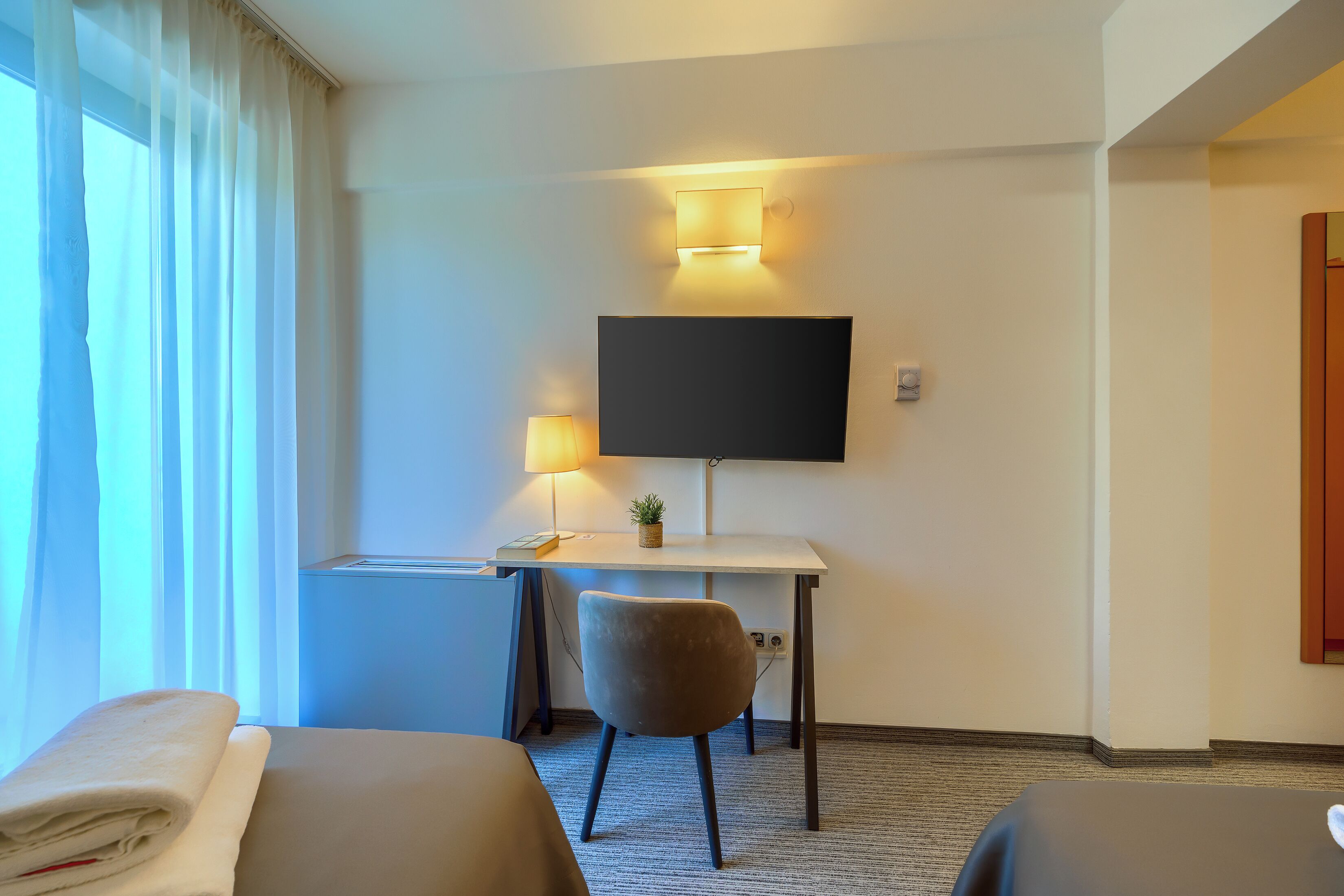 Una habitación de hotel con cama, escritorio, silla, televisión y ventana con cortinas.