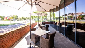 An outdoor patio area with wicker furniture and large umbrellas, overlooking a street and greenery.