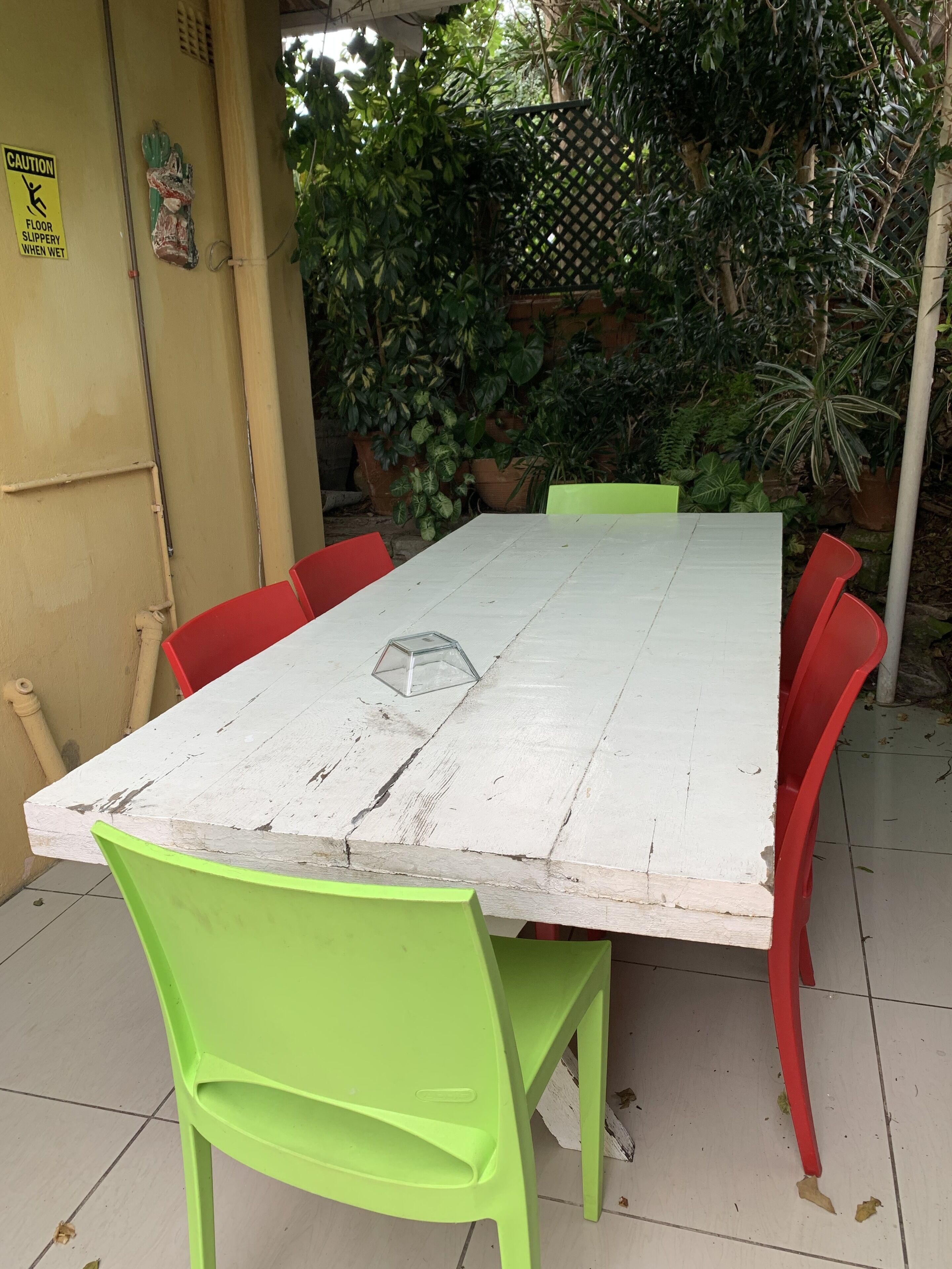 A white wooden table with red and green chairs, surrounded by potted plants and a caution sign on a yellow wall.