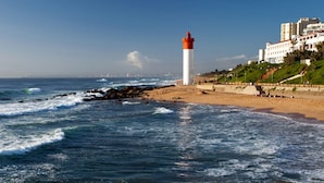 Paisaje costero con un faro, playa y edificios al fondo.