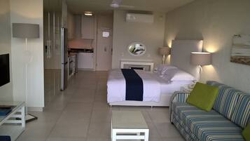 A hotel room with a bed, a sofa, a small table, a TV, a kitchen area, and a bathroom.