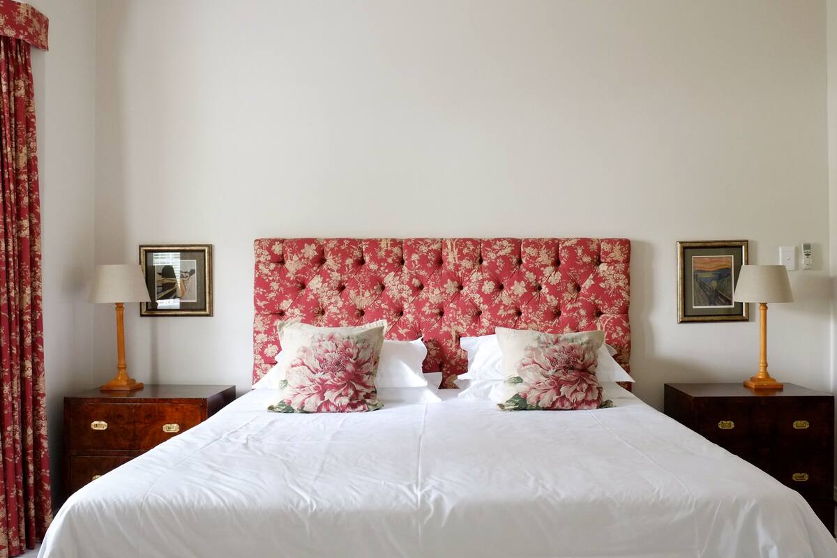 A bedroom with a floral headboard, two bedside tables with lamps, a bed with white bedding, and a red curtain.