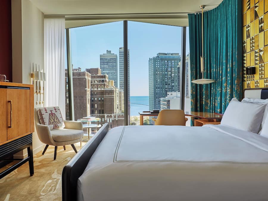 A hotel room with a large bed, a wooden cabinet, a chair, and a view of the city skyline.