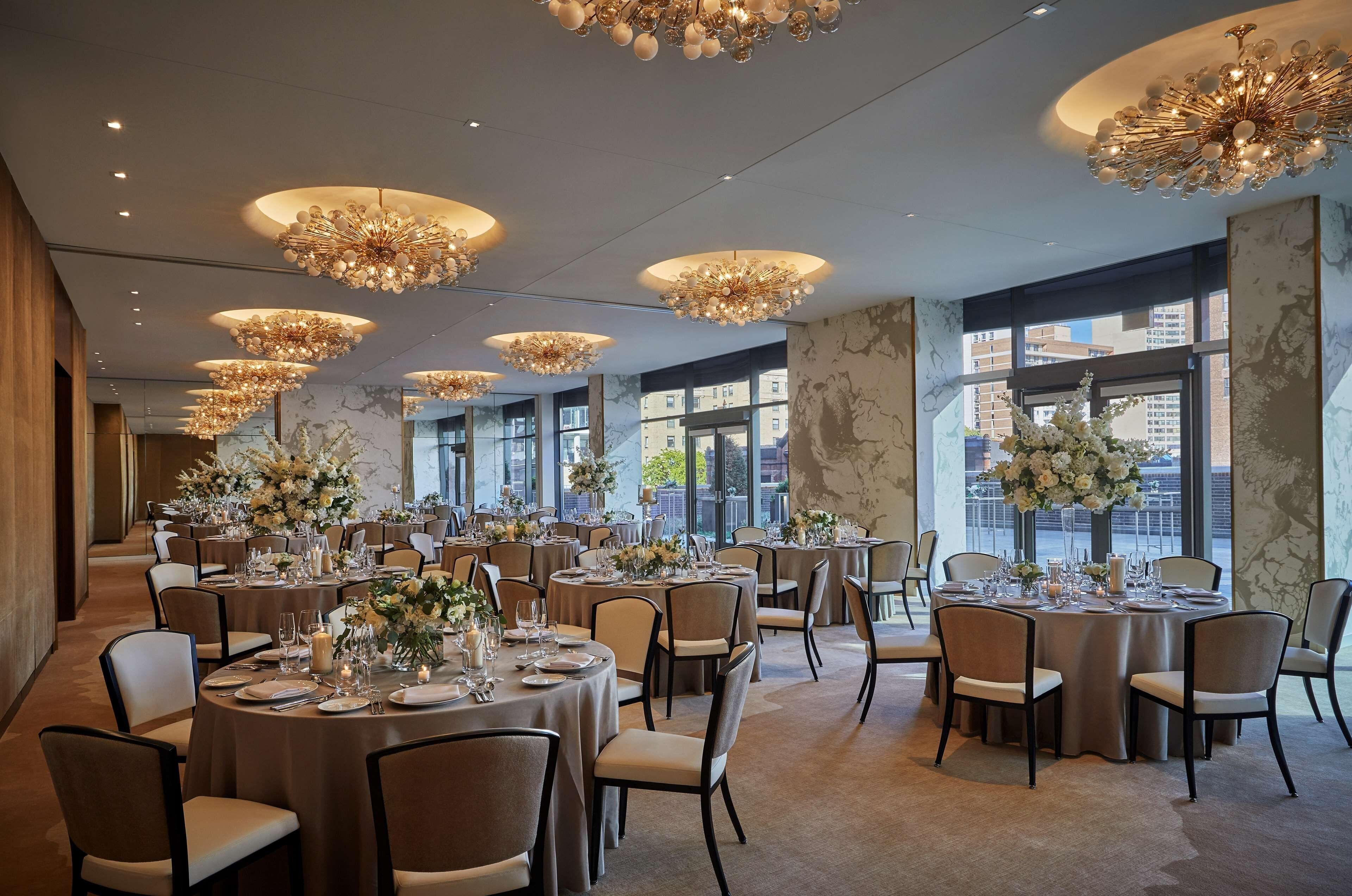 A spacious banquet hall with multiple round tables set for dining, elegant chandeliers, and large windows offering city views.
