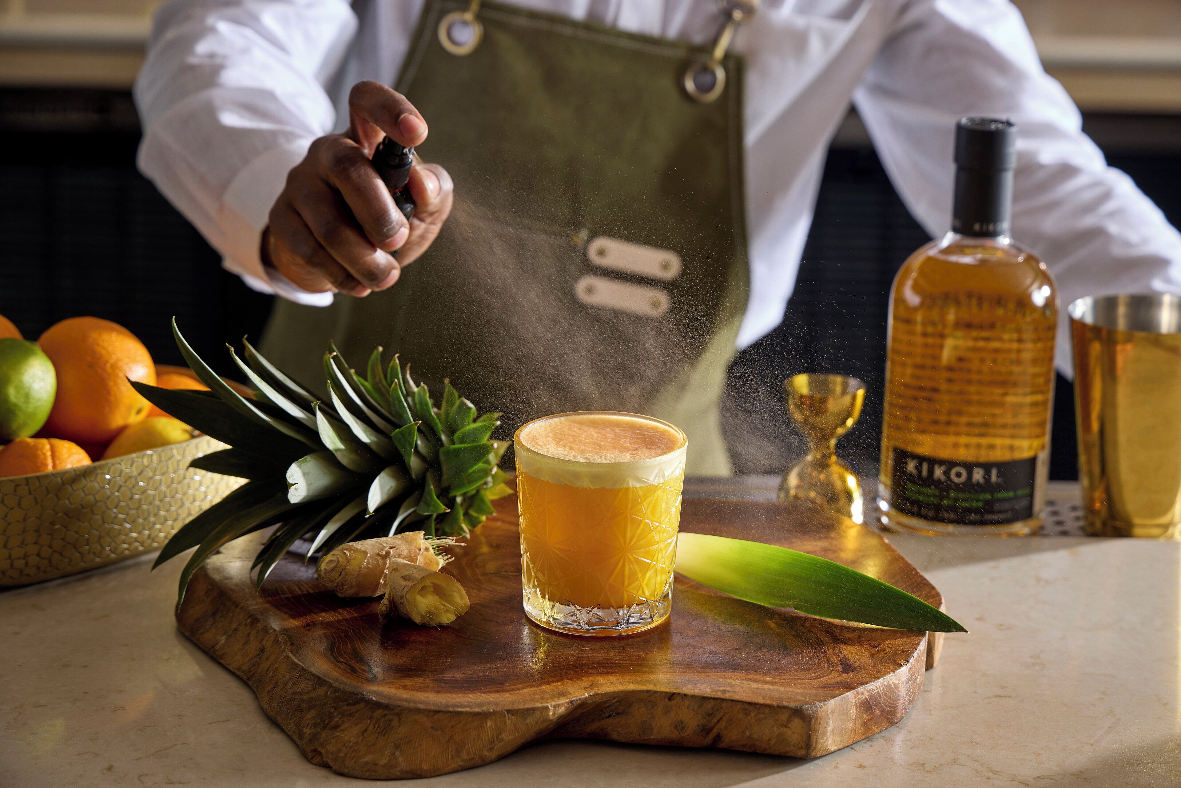 A bartender preparing a drink with a lime, pineapple, and ginger on a wooden board, with a bottle of KIKORI in the background.