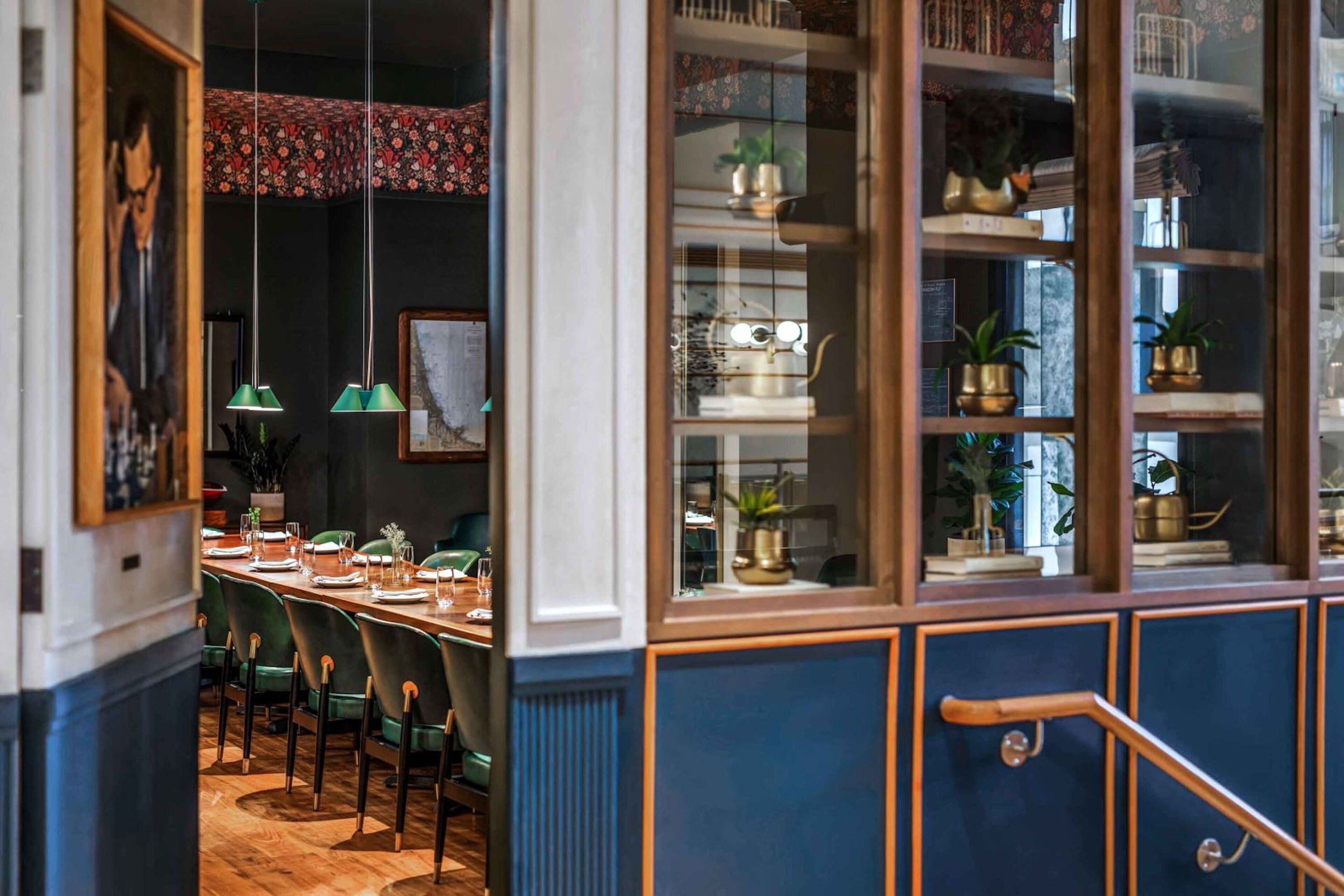 A dining area with a long table set for four, green chairs, hanging green pendant lights, and a blue and white cabinet with glass doors displaying decorative items.