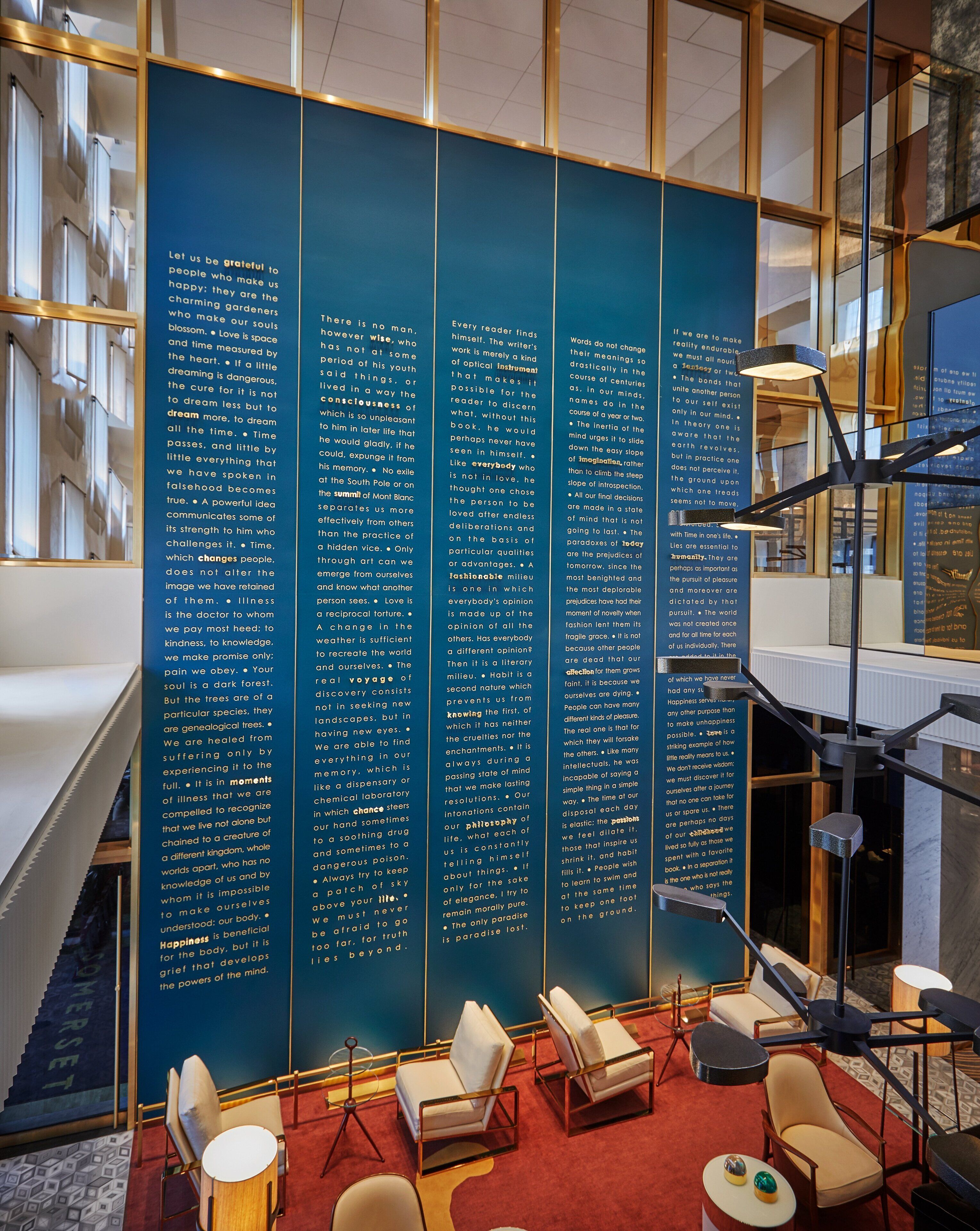 A modern interior with a large blue wall displaying text, seating area with chairs and tables, and a staircase with glass railings.